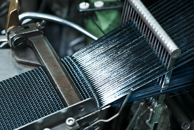 Detailaufnahme einer industriellen Fertigungsanlage, in der zahlreiche schwarze Metall- oder Kunststoffdrähte präzise geführt und zu einem dichten Geflecht verarbeitet werden.